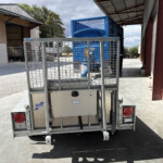 Rear view of a metal utility trailer loaded with a blue, cage-like container in an industrial yardyard setup