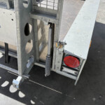 Close-up of a metal trailer frame showing a hydraulic jack and red taillight, with riveted silver panels and a textured ground surface in sunlight.