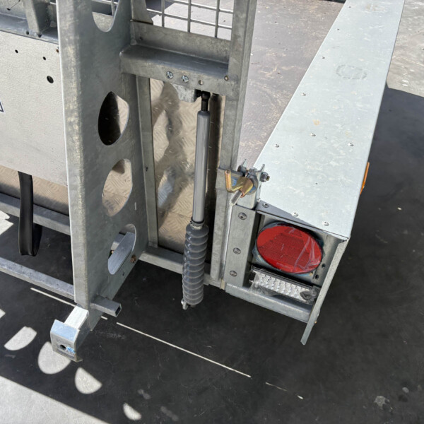 Close-up of a metal trailer frame showing a hydraulic jack and red taillight, with riveted silver panels and a textured ground surface in sunlight.