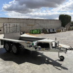 Flatbed trailer parked on a concrete lot beside a tall concrete wall, with metal bins and gravel piles in the background under a cloudy sky.