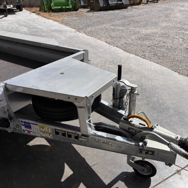 Close-up of a metal utility trailer hitch and ramp on a concrete surface with a gravel lot in the background.