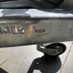 Industrial metal surface with stamped numbers and a GX84 barcode sticker, shadows of a person cast nearby, and a wheel visible below.