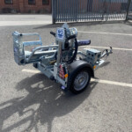 Trailer hitch and utility trailer frame in a parking lot, with a metal gate in the background and a brick building.