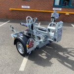 Metal trailer in a parking lot beside a brick building, with a bright yellow safety rail and 'Visitor Parking' signs in the background