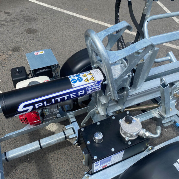 Trailer-mounted gas-powered log splitter with a blue metal frame and safety stickers, parked on asphalt surface.