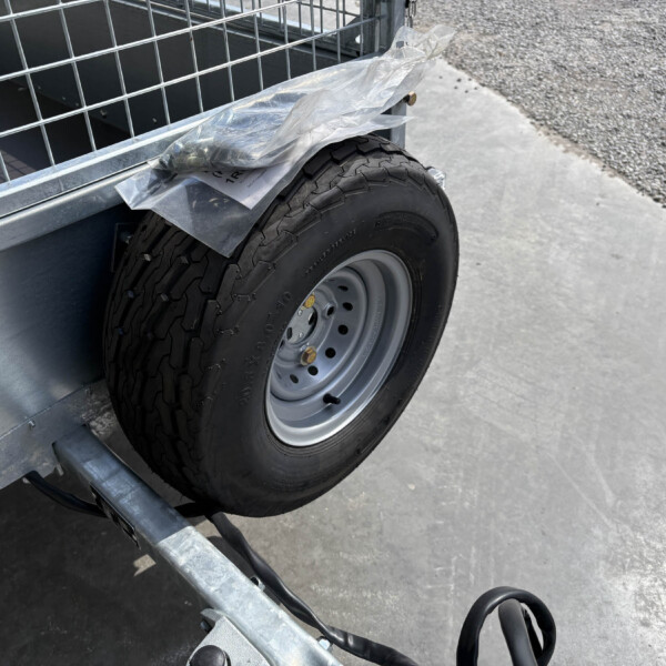 Trailer wheel mounted on a metal frame, with a plastic bag resting on the fender and gravel ground in the background.