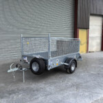 Silver metal utility trailer with mesh sides and a tow hitch, parked in front of a large corrugated metal roll-up door in an industrial area.