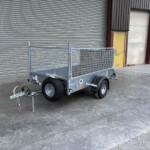 Metal utility trailer with mesh sides and hitch, parked on a concrete floor in front of a large gray industrial roller door.