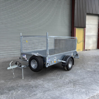 Silver metal utility trailer with mesh sides and a tow hitch, parked in front of a large corrugated metal roll-up door in an industrial area.