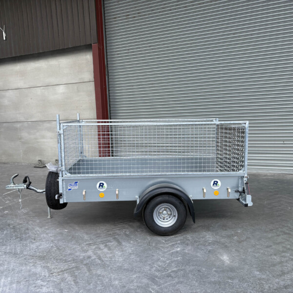 Metal utility trailer with mesh sides and a ramp, parked in front of a large corrugated metal shutter at a warehouse.