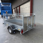 Blue industrial trailer with a tall, cage-like sides unit parked in a gravel lot beside a corrugated metal building, awaiting transport.