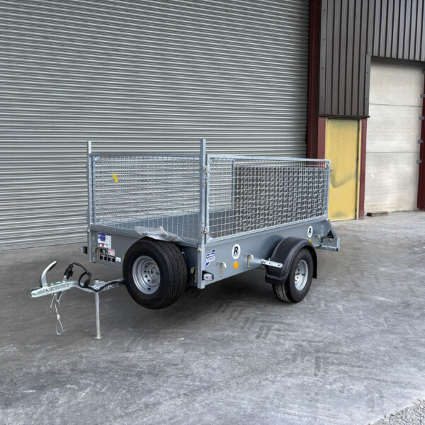 Silver metal utility trailer with mesh sides and a tow hitch, parked in front of a large corrugated metal roll-up door in an industrial area.