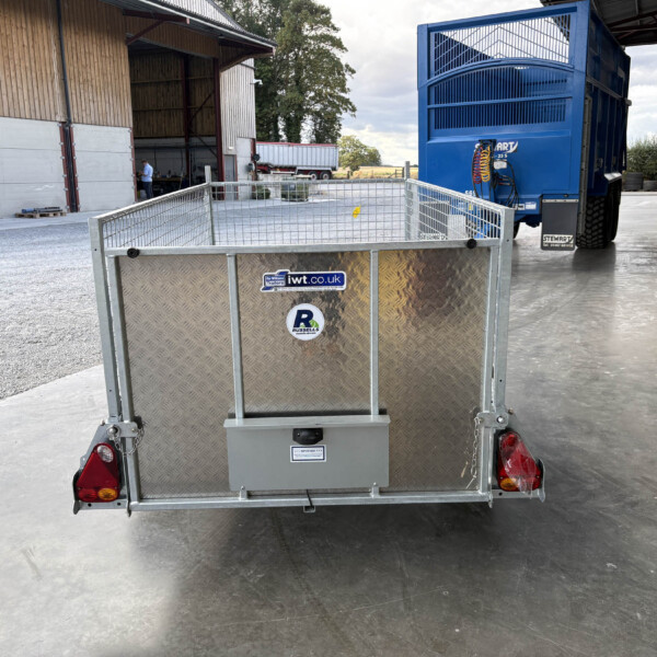 Rear view of a diamond-plate trailer with red taillights, hitched to a blue truck inside a loading yard.