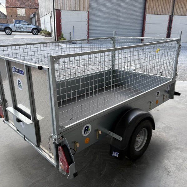 Metal mesh-sided utility trailer in a concrete yard near warehouses, with a ramp and dual wheels visible at the back.