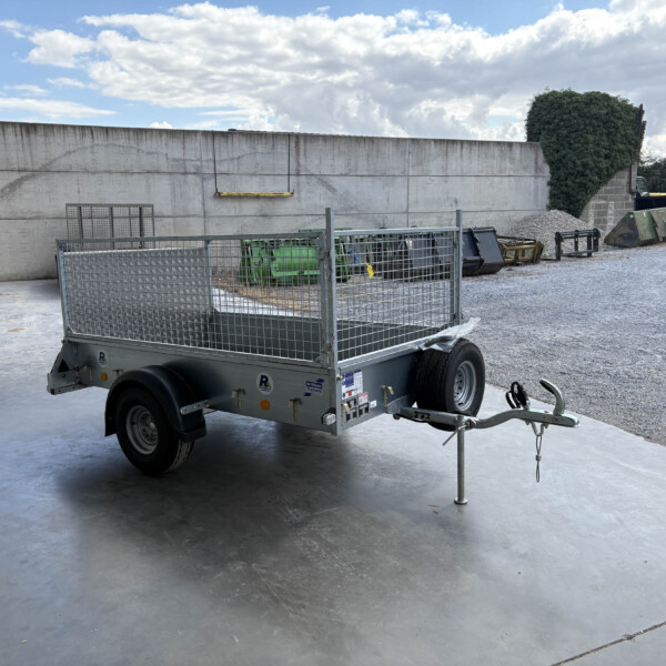Metal mesh-sided utility trailer in a gravel yard beside a tall concrete wall and gravel pile in the background.
