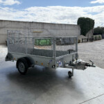 Open metal utility trailer with mesh sides parked in a gravel lot beside a tall concrete wall and scattered supplies under a blue sky with clouds.