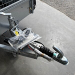Close-up of a trailer hitch and jack on a gray utility trailer, with a plastic bag around a wiring harness on a concrete surface