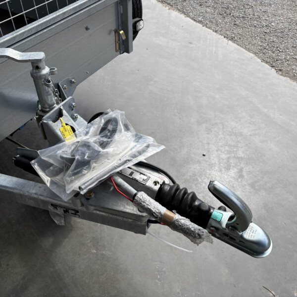 Close-up of a trailer hitch and jack on a gray utility trailer, with a plastic bag around a wiring harness on a concrete surface