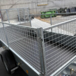 Galvanized metal mesh utility trailer with a gated side, parked in an industrial yard with dumpsters in the background.