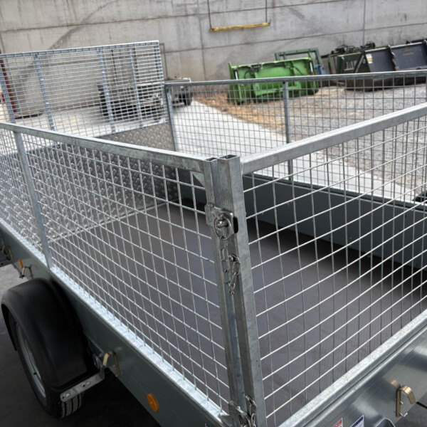 Galvanized metal mesh utility trailer with a gated side, parked in an industrial yard with dumpsters in the background.