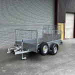 Dual-axle metal utility trailer with mesh sides parked in front of a gray corrugated roll-up door.