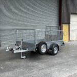 Metal utility trailer with mesh sides in front of a large gray corrugated roll-up door in an industrial loading area.