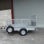 Silver metal utility trailer with mesh sides and two axles parked on a concrete floor in front of a large corrugated metal roll-up door.