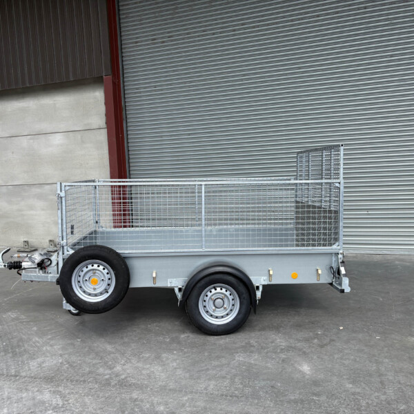 Silver metal utility trailer with mesh sides and two axles parked on a concrete floor in front of a large corrugated metal roll-up door.