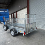 Blue double-axle livestock trailer with grated sides parked beside a corrugated metal wall and large roll-up door in an industrial yard.