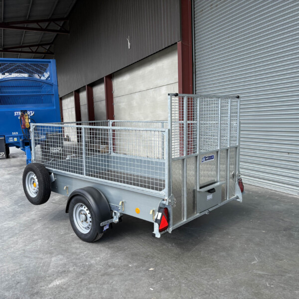 Blue double-axle livestock trailer with grated sides parked beside a corrugated metal wall and large roll-up door in an industrial yard.