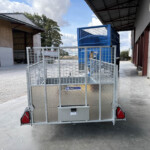 Rear view of a metal utility trailer with a diamond-plate bed and blue mesh enclosure inside an industrial loading bay.
