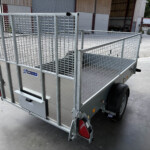 Rear three-quarter view of a galvanized metal utility trailer with mesh sides and red tail lights in an industrial yard.