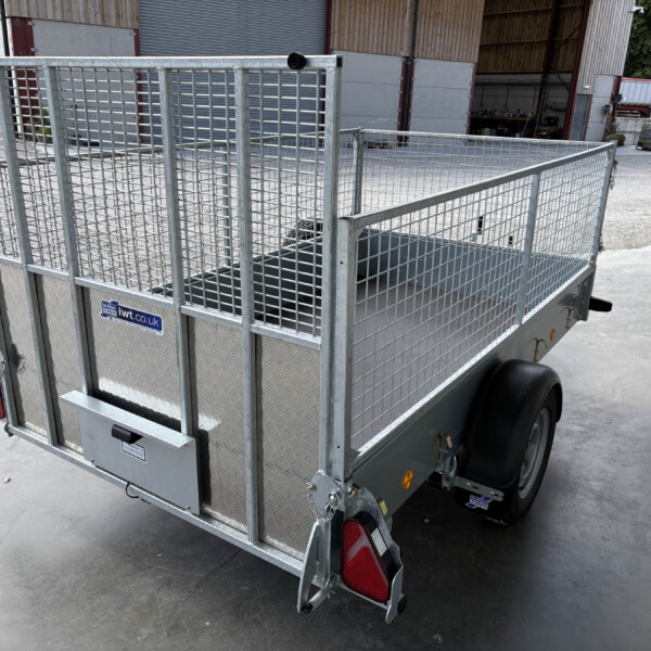 Rear three-quarter view of a galvanized metal utility trailer with mesh sides and red tail lights in an industrial yard.