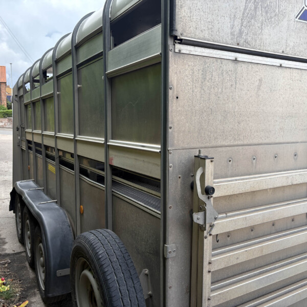 Close-up of a large gray livestock trailer with metal panels, wheels, and a partially open side door on a street edge.