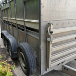 Side view of a weathered metal trailer with vertical panels and twin wheels, parked beside a concrete surface with green plants in the foreground and a brick building in the background.