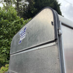 Close-up of a weathered metal trailer with a blue-and-white logo sticker, green hedges, and cloudy sky in the background.