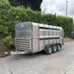 Metal livestock trailer parked on a street beside a tall green hedge, with a streetlight pole and brick building in the background.