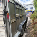 Green livestock trailer parked along a path beside a hedge and a building with Russell's sign in the background, showing wheels and rear details.