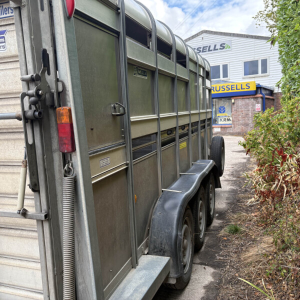 Green livestock trailer parked along a path beside a hedge and a building with Russell's sign in the background, showing wheels and rear details.