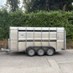 Silver metal livestock trailer parked on an asphalt surface with a dense green hedge and trees in the background behind it.”,“A large aluminum trailer with multiple wheels sits in front of a tall green hedge.”,“Side view of a silver trailer with vent openings, positioned before a thick hedge and trees.”,“Livestock trailer on a parking area with a green hedge and trees behind it.”