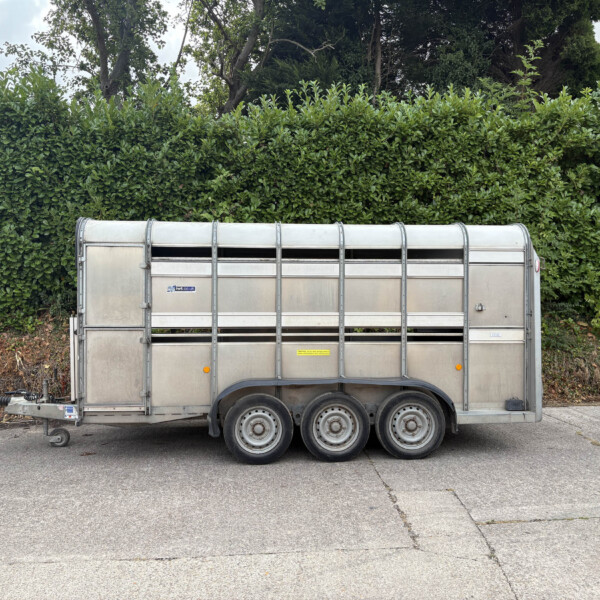 Silver metal livestock trailer parked on an asphalt surface with a dense green hedge and trees in the background behind it.”,“A large aluminum trailer with multiple wheels sits in front of a tall green hedge.”,“Side view of a silver trailer with vent openings, positioned before a thick hedge and trees.”,“Livestock trailer on a parking area with a green hedge and trees behind it.”