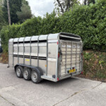 Metal livestock trailer parked in a paved lot next to a tall green hedge and a white building with blue-and-yellow signage visible nearby