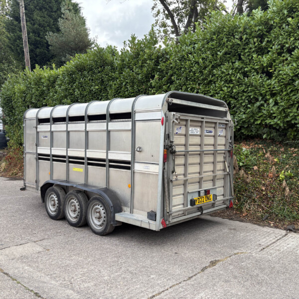 Metal livestock trailer parked in a paved lot next to a tall green hedge and a white building with blue-and-yellow signage visible nearby