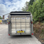 Rear view of a metal trailer with its tailgate closed, displaying the UK plate XD22 ONG and various stickers, in a paved lot with a hedge on the right and a building in the background on a cloudy day.