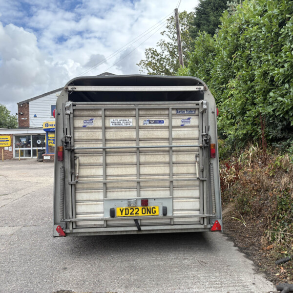 Rear view of a metal trailer with its tailgate closed, displaying the UK plate XD22 ONG and various stickers, in a paved lot with a hedge on the right and a building in the background on a cloudy day.