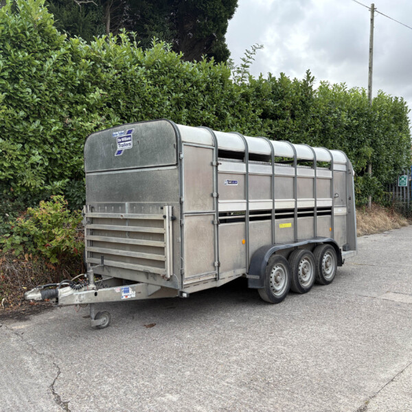 Metal livestock trailer parked on a street beside a tall green hedge, with a streetlight pole and brick building in the background.