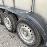Close-up of tandem trailer wheels with a long curved black fender and a gray trailer wall behind them, on an asphalt surface.