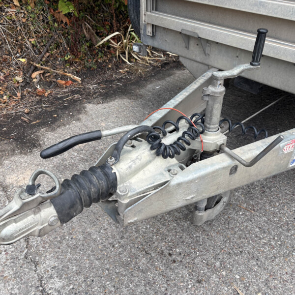 Close-up of a trailer hitch coupling with coiled electrical cables on a gravel surface near a plant-filled area.