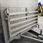 Close-up of a corrugated metal trailer side with a hitch and wheel, with yellow construction machinery in the background.