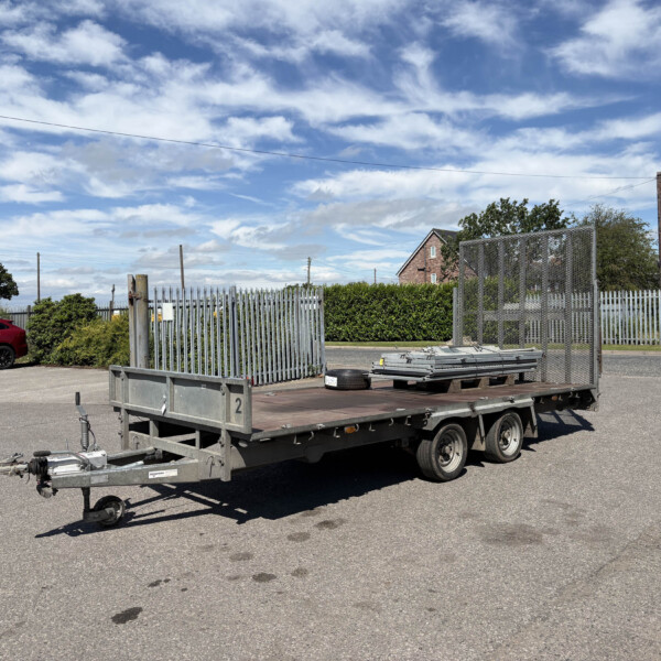 Flatbed trailer with side rails in a parking lot, a red car visible on the left and a chain-link fence in the background under a blue sky with clouds.